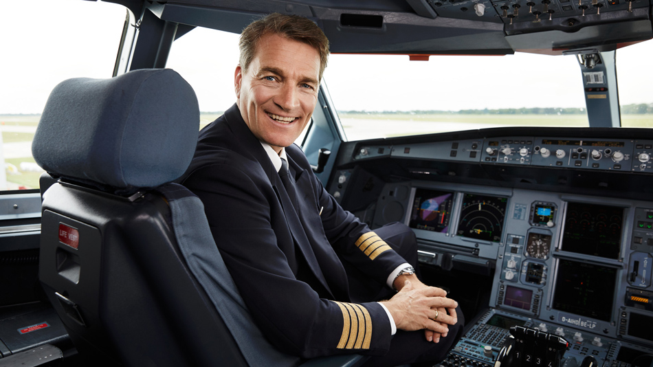 An aircraft captain sits in a cockpit and smilingly looks into the camera